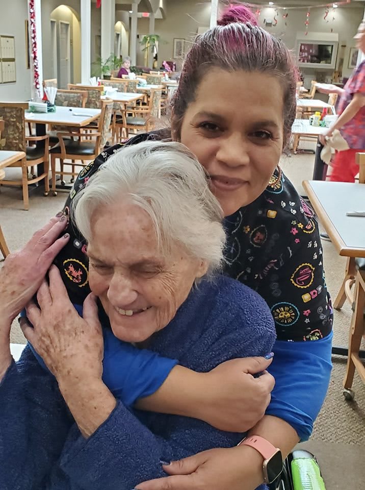 A caregiver warmly embraces an elderly woman, both smiling.