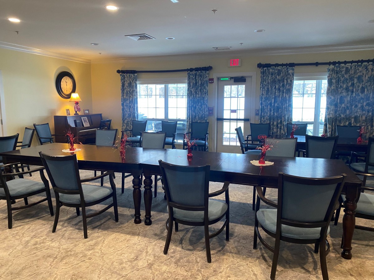 A spacious dining room with large tables and several chairs, featuring bright windows and decorative curtains.