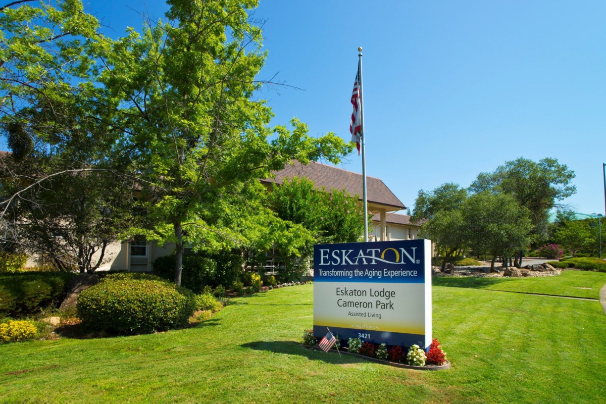 A sign for Eskaton Village Carmichael is displayed on a well-maintained lawn, accompanied by trees and an American flag on a sunny day.