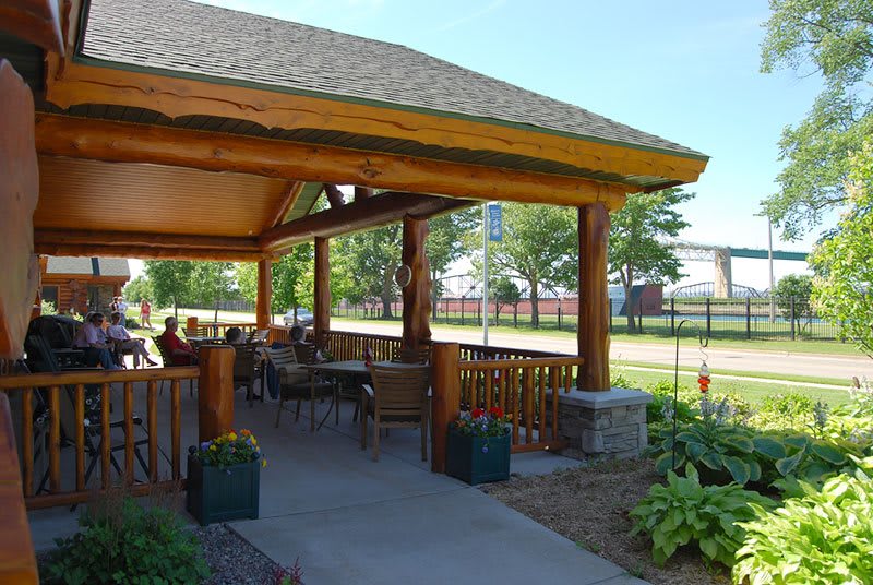 A cozy outdoor patio with wooden beams and seating, surrounded by a garden.