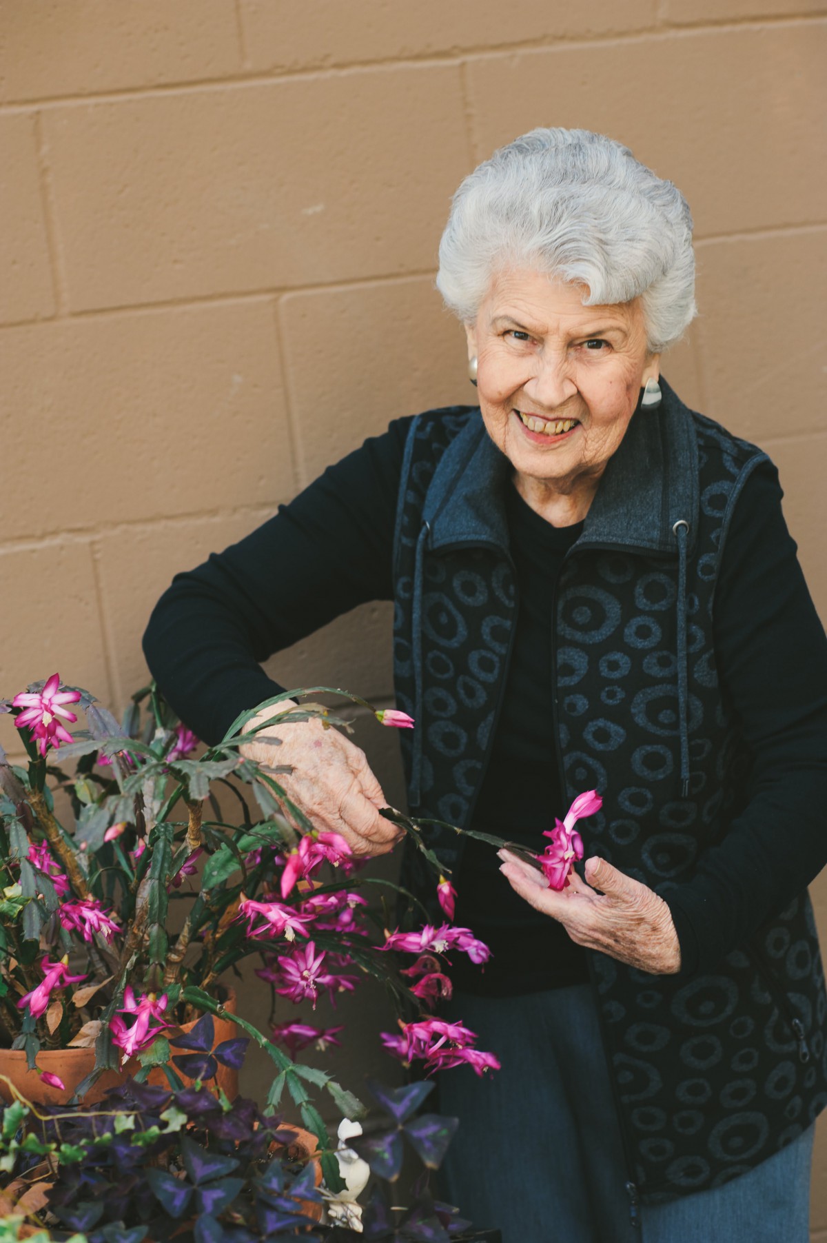 An elderly woman smiling while tending to a vibrant flowering plant indoors.