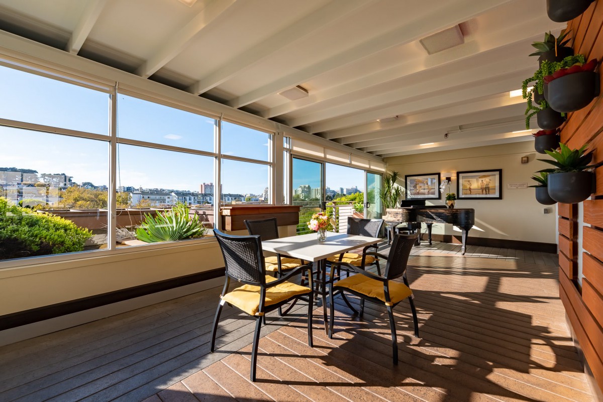 A modern dining area with large windows allowing natural sunlight, featuring a table set with chairs and a piano in the corner.