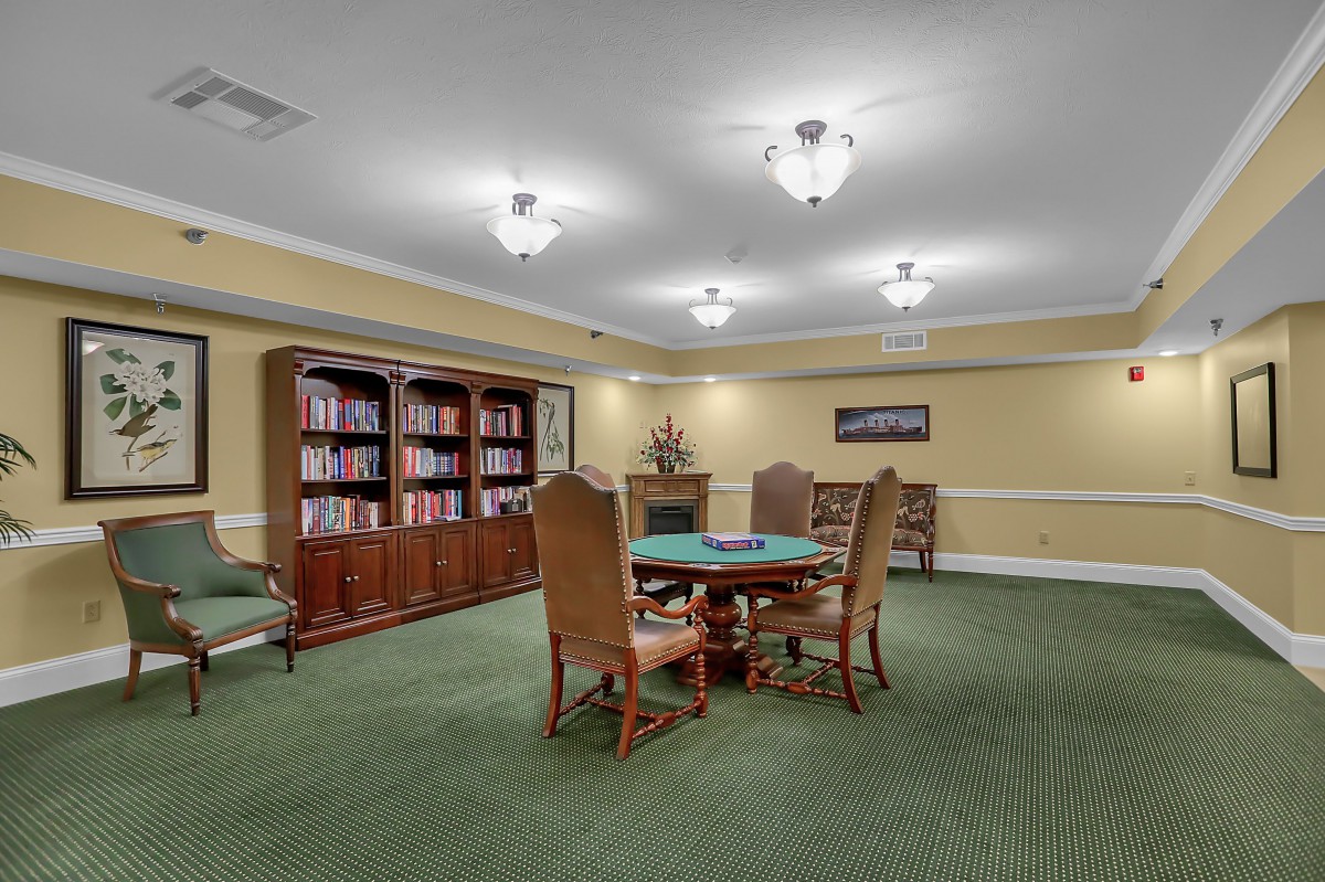 A cozy library room with bookshelves, a round table, and chairs on a carpeted floor.
