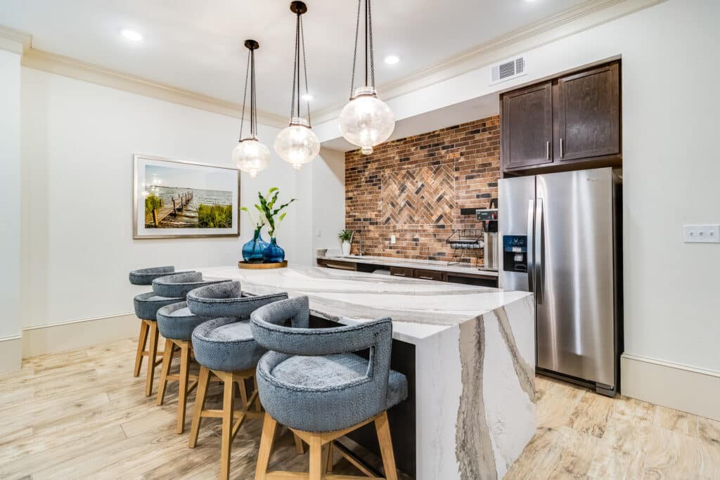 A modern kitchen featuring a large island with bar stools, pendant lighting, a stone backsplash, and stainless steel appliances.