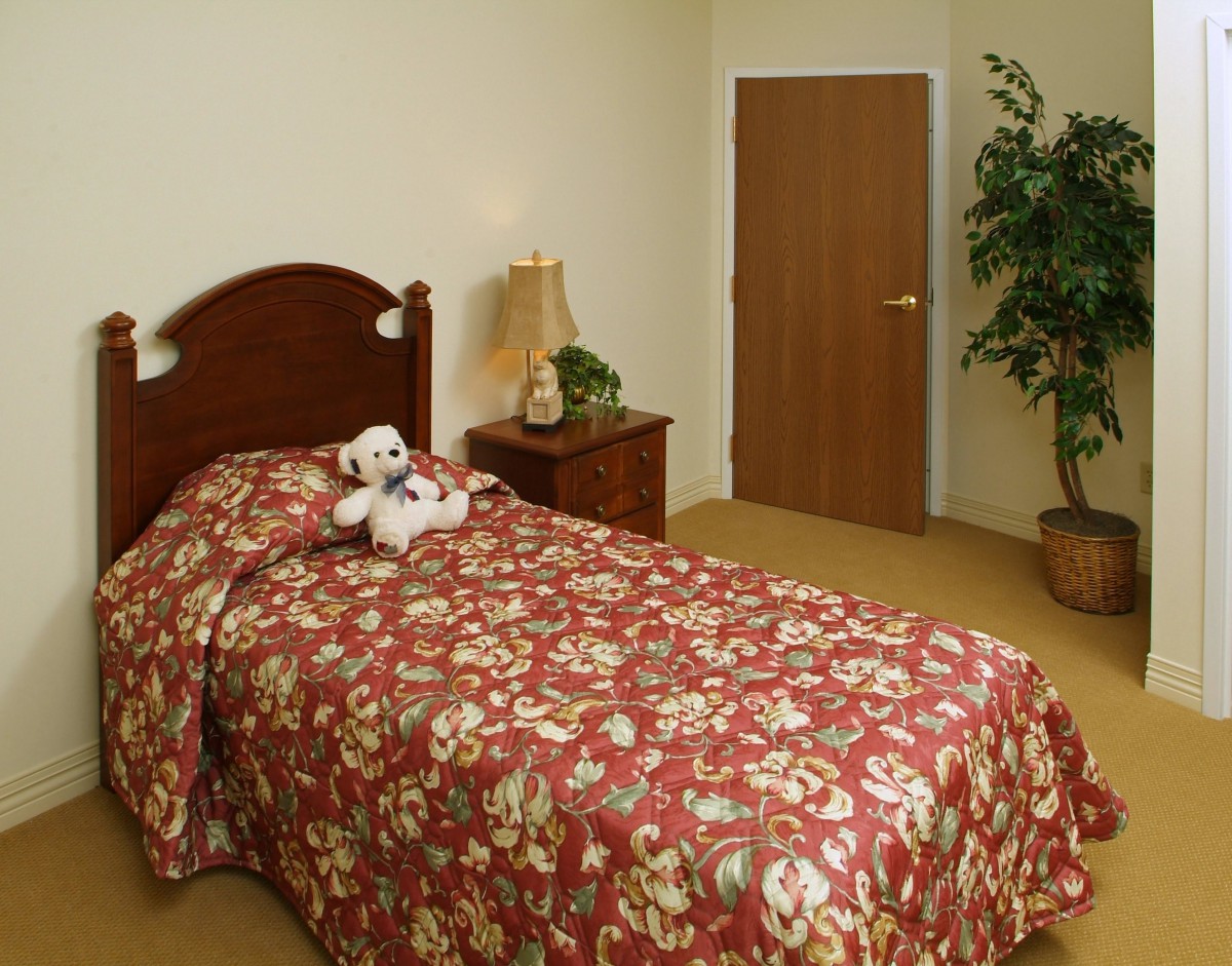 A cozy bedroom features a single bed with a floral bedspread, adorned with a teddy bear, alongside a nightstand and a lamp. A tall plant stands in the corner near the door.