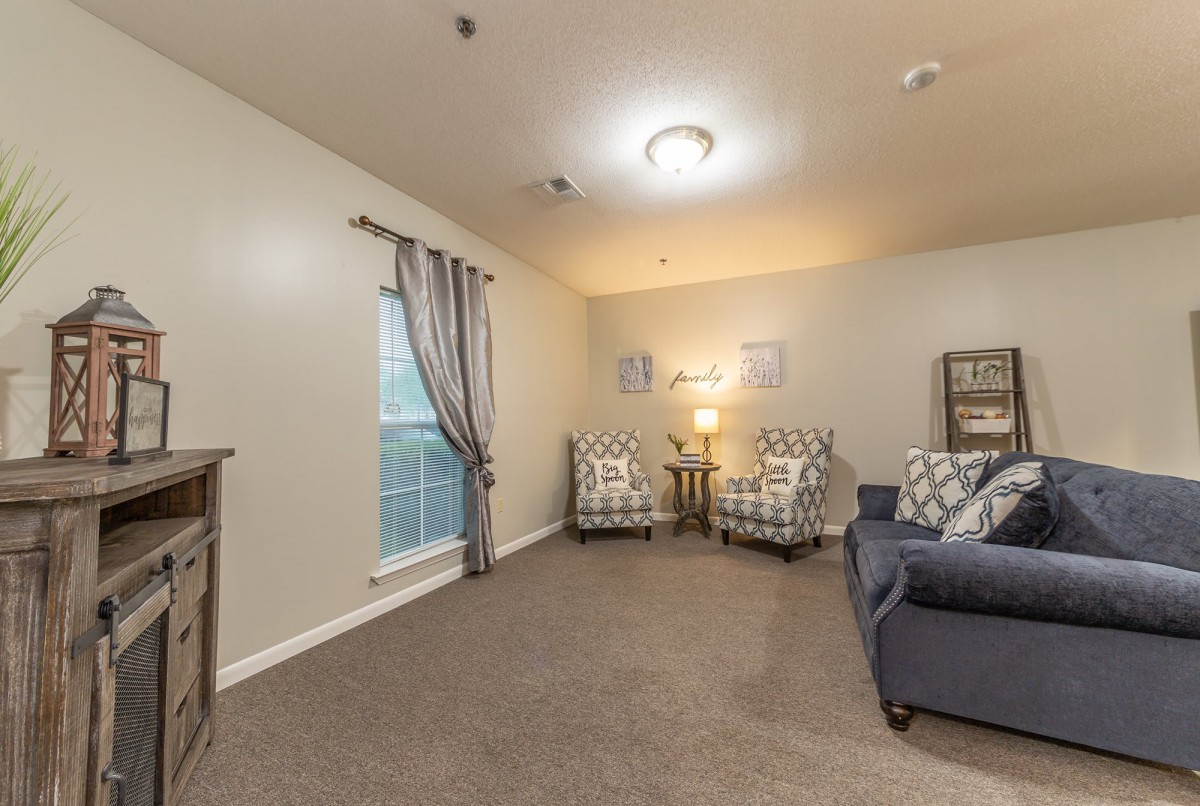 A cozy living room with a sofa, two patterned armchairs, a lamp, and curtains by the window.