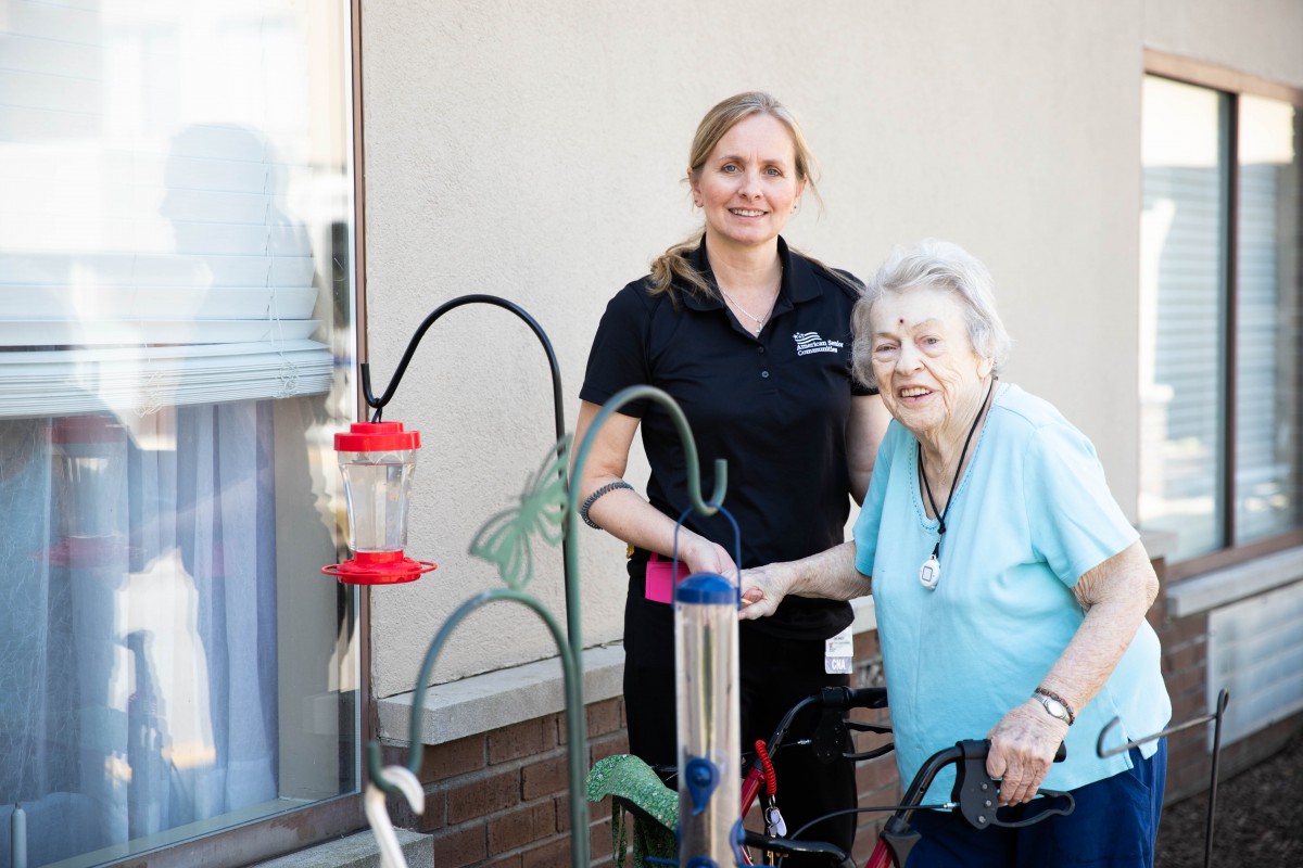 A caregiver assisting an elderly woman with bird feeders outside.