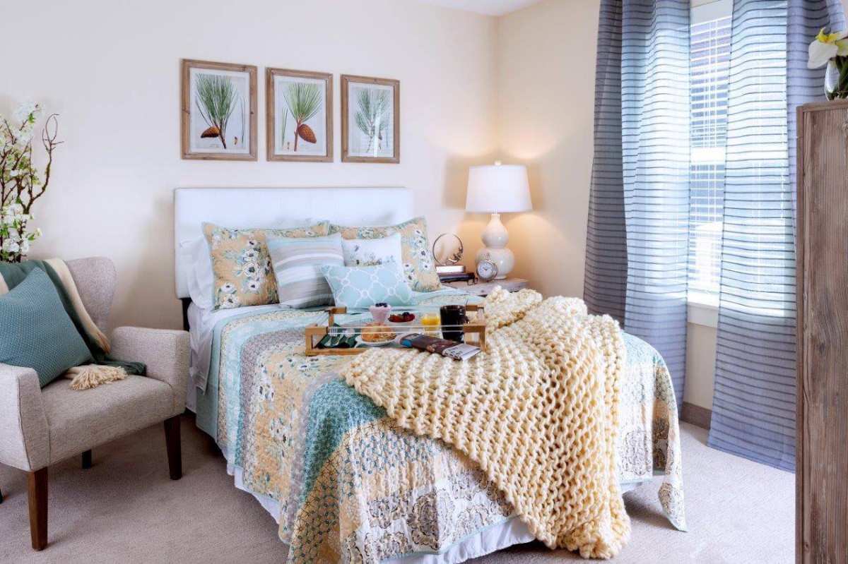 A cozy bedroom featuring a neatly made bed with patterned pillows and a chunky knit blanket, complemented by soft lighting from a table lamp and artwork above the headboard.