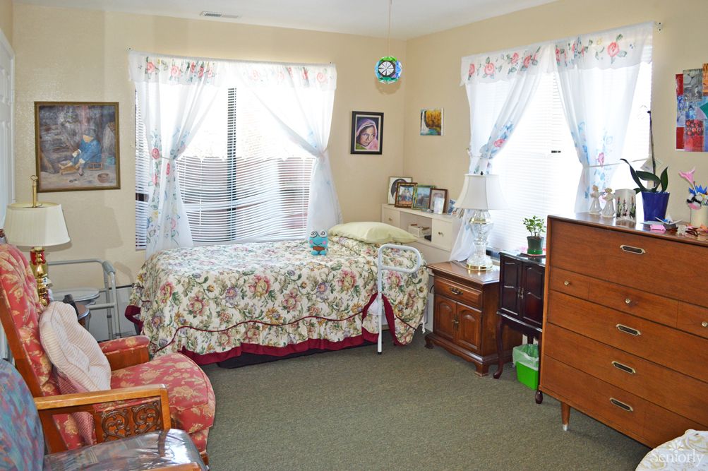 A cozy bedroom with a bed, dresser, and chair, featuring floral bedding and sheer curtains.