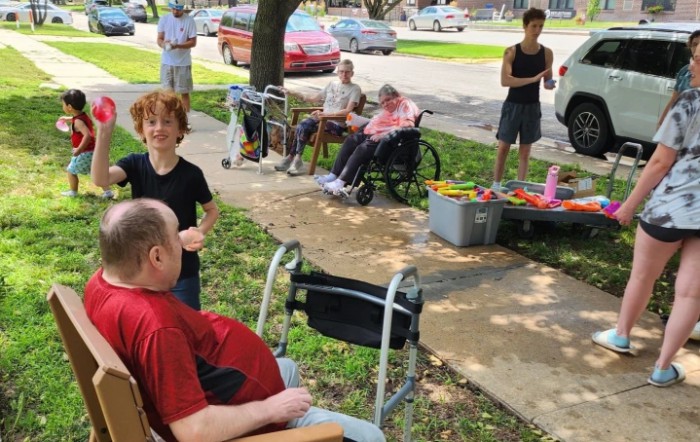 A group of people enjoying an outdoor community event, with some individuals seated and others standing around a table on a sunny day.