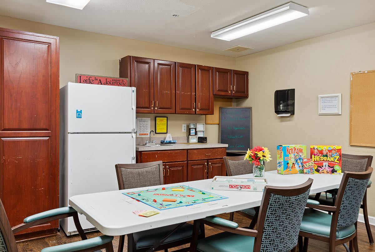 A cozy kitchen area with a table set for games and flowers, surrounded by cabinets and appliances.