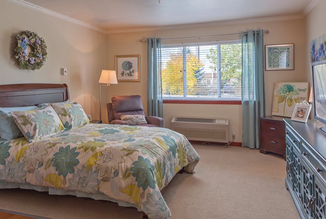 A cozy bedroom interior with a bed, armchair, lamp, and large window.