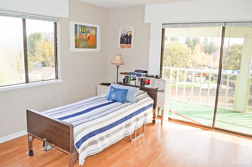 A cozy bedroom with a single bed featuring striped bedding, a small bookshelf, and a lamp by the window.