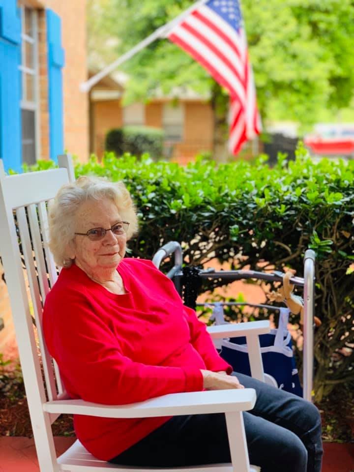 An elderly woman relaxes on a rocking chair on the patio, with an American flag in the background.