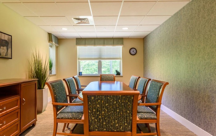 A well-lit conference room with a wooden table, surrounded by upholstered chairs, featuring a window and indoor plant.