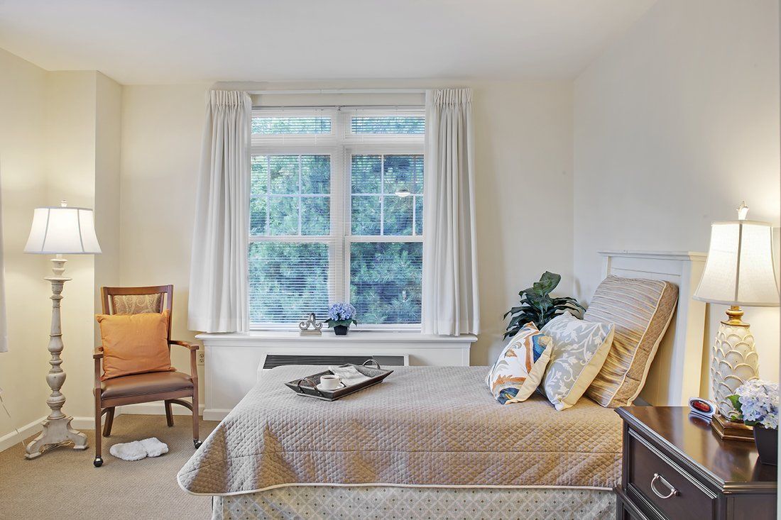 A cozy bedroom with a neatly made bed, a wooden chair beside a large window letting in natural light.