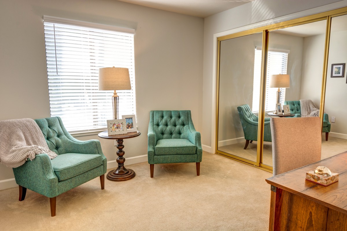 A cozy living room with two teal armchairs, a wooden side table, and a large mirror reflecting light from the window.