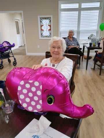 An elderly woman smiling while holding a pink elephant balloon indoors.