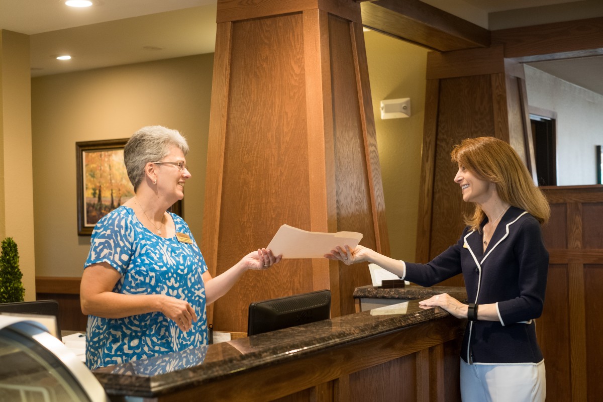 A receptionist handing a document to a woman at a counter.