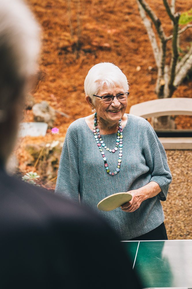 An elderly woman smiles warmly outdoors, holding an object in her hand. She is wearing glasses and a beaded necklace, with autumn leaves in the background.