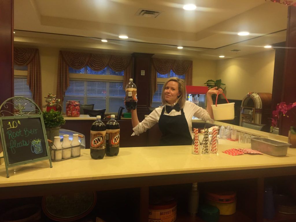A person standing behind a soda fountain counter, smiling and holding a bottle.