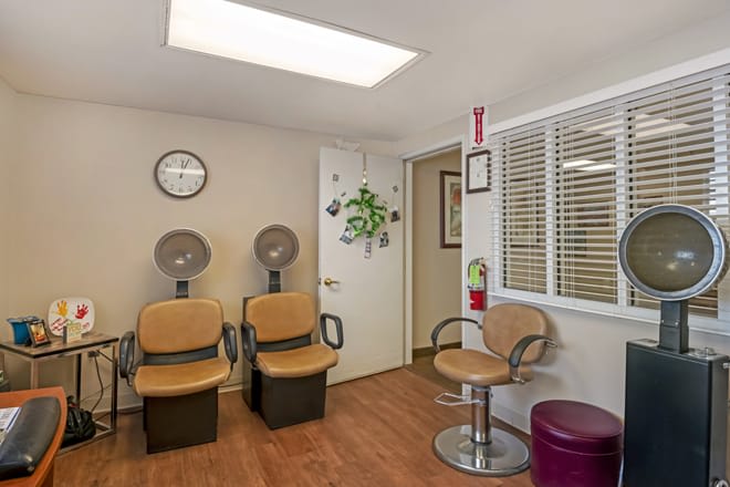 A cozy salon waiting area with chairs and hair dryers, featuring a wall clock and wooden flooring.