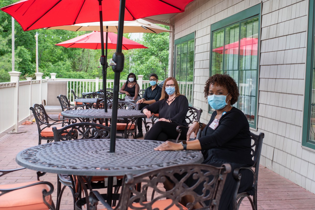A group of individuals wearing masks are seated at an outdoor patio, practicing social distancing under red umbrellas.