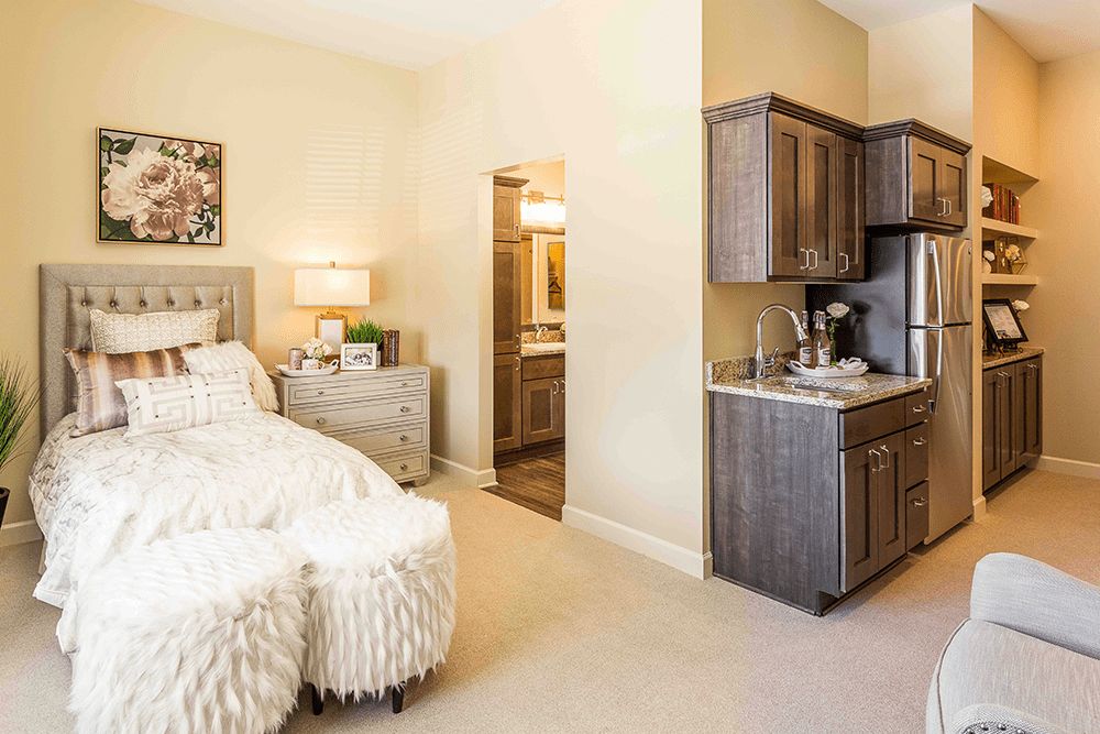 A cozy bedroom with a kitchenette, featuring a plush bed, side table, lamp, and decorative elements.