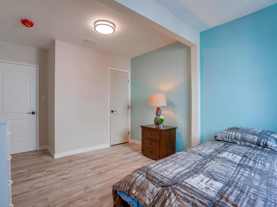 A cozy bedroom with a blue accent wall, featuring a bed with patterned bedding, a nightstand with a decorative lamp, and wood flooring.
