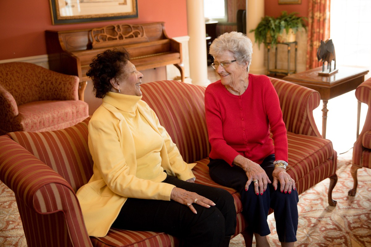 Two elderly women sitting on a couch and engaging in a lively conversation.