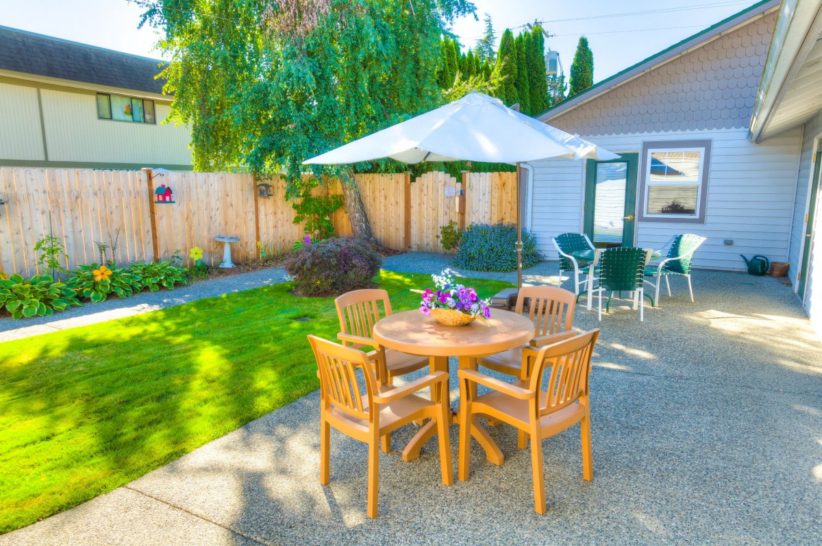 A cozy backyard patio with a wooden table and chairs under a large umbrella, surrounded by a well-maintained garden.