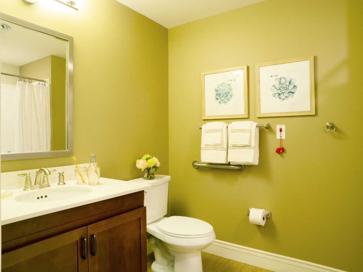 A neatly decorated bathroom with a yellow-green wall, featuring a toilet, sink, towels, mirror, and framed artwork.