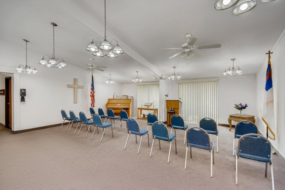 An empty chapel with rows of blue chairs, a pulpit, an organ, and a cross on the wall.