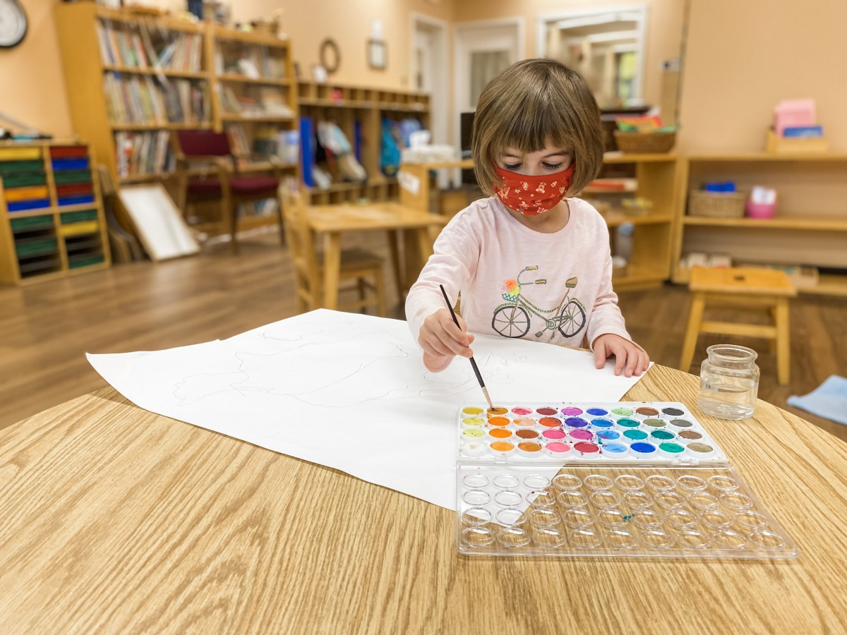 A young child wearing a mask is engaged in painting with watercolors at a table indoors.