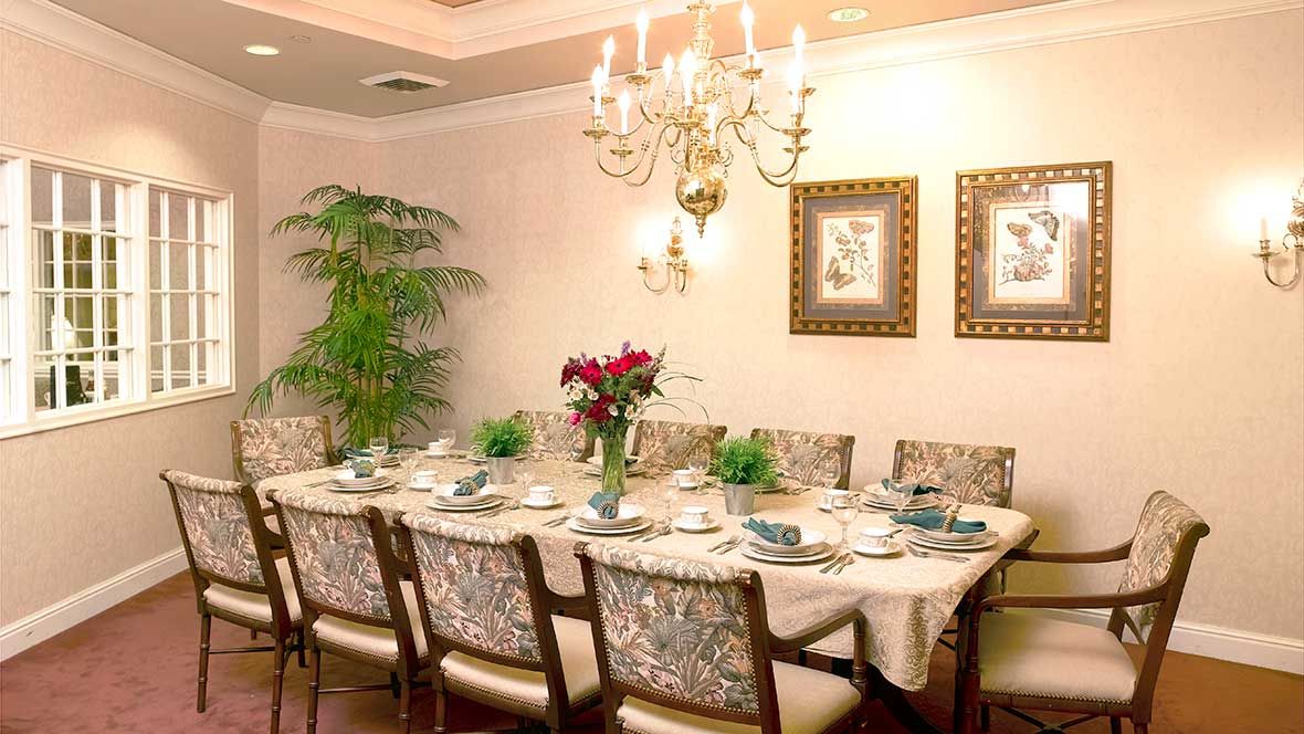 An elegantly decorated dining room with a long table set for guests, featuring a chandelier and floral centerpiece.