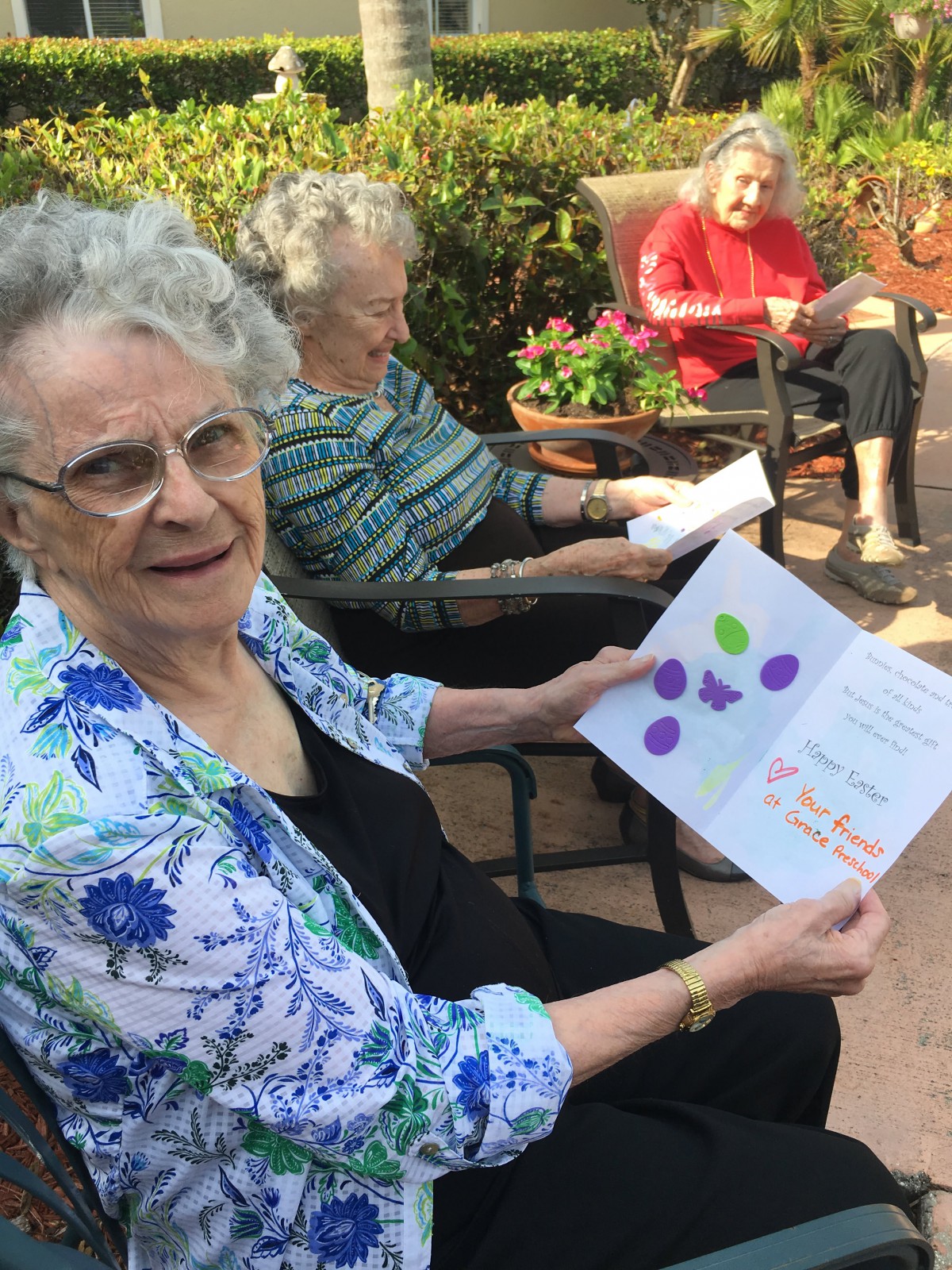 An elderly woman seated outdoors in a garden, holding and showing a card while others sit nearby.
