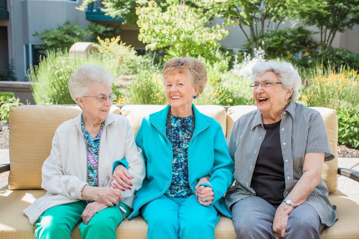 Three elderly women enjoying each other's company while sitting on a bench outdoors.
