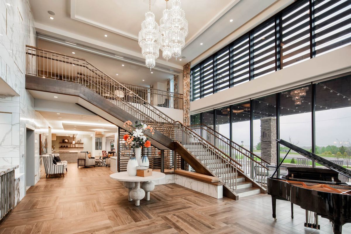A luxurious lobby featuring a grand staircase, elegant chandelier, and a grand piano, with large windows providing ample natural light.