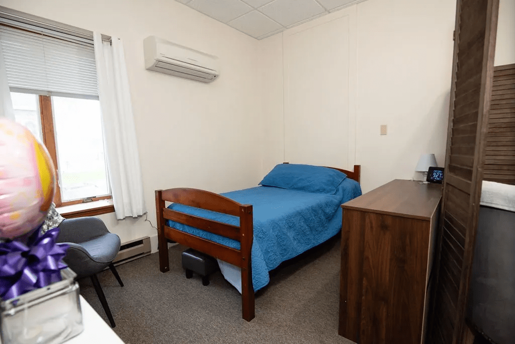 A small bedroom with a single bed covered in blue bedding, a wooden dresser, a chair by the window, and an air conditioning unit on the wall.