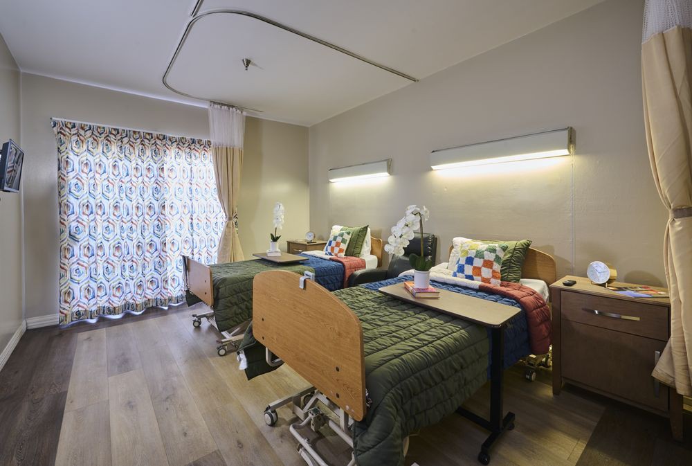 A hospital room with two patient beds equipped with bedside tables and overhead lighting.