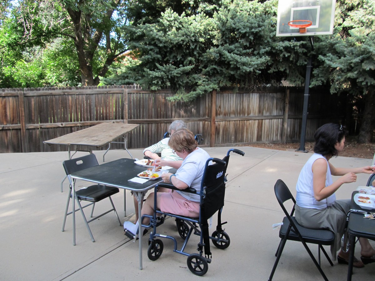 A person in a wheelchair sits at a table outdoors, next to another individual with a basketball hoop in the background.