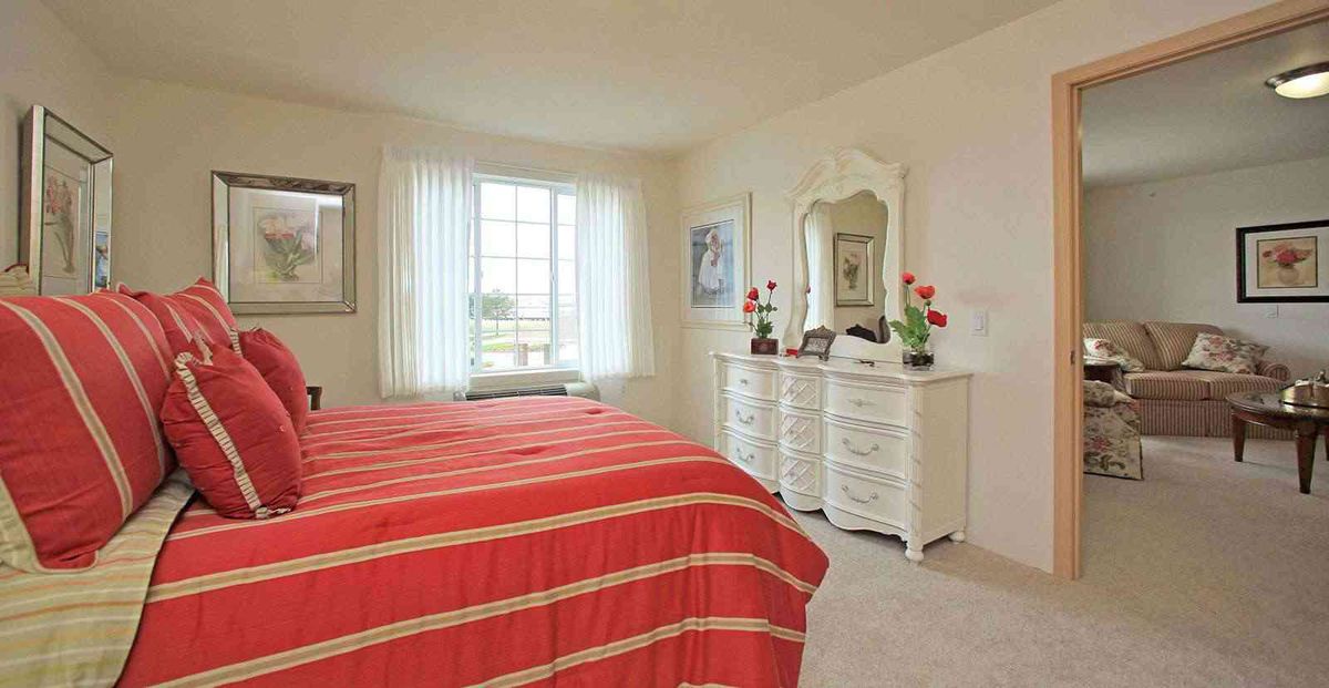 A cozy bedroom with a red bedspread, a dresser with a mirror, and natural light coming through a window.