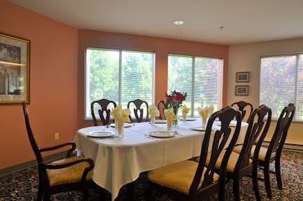 A dining room with a table set for six, featuring floral arrangements by the windows.
