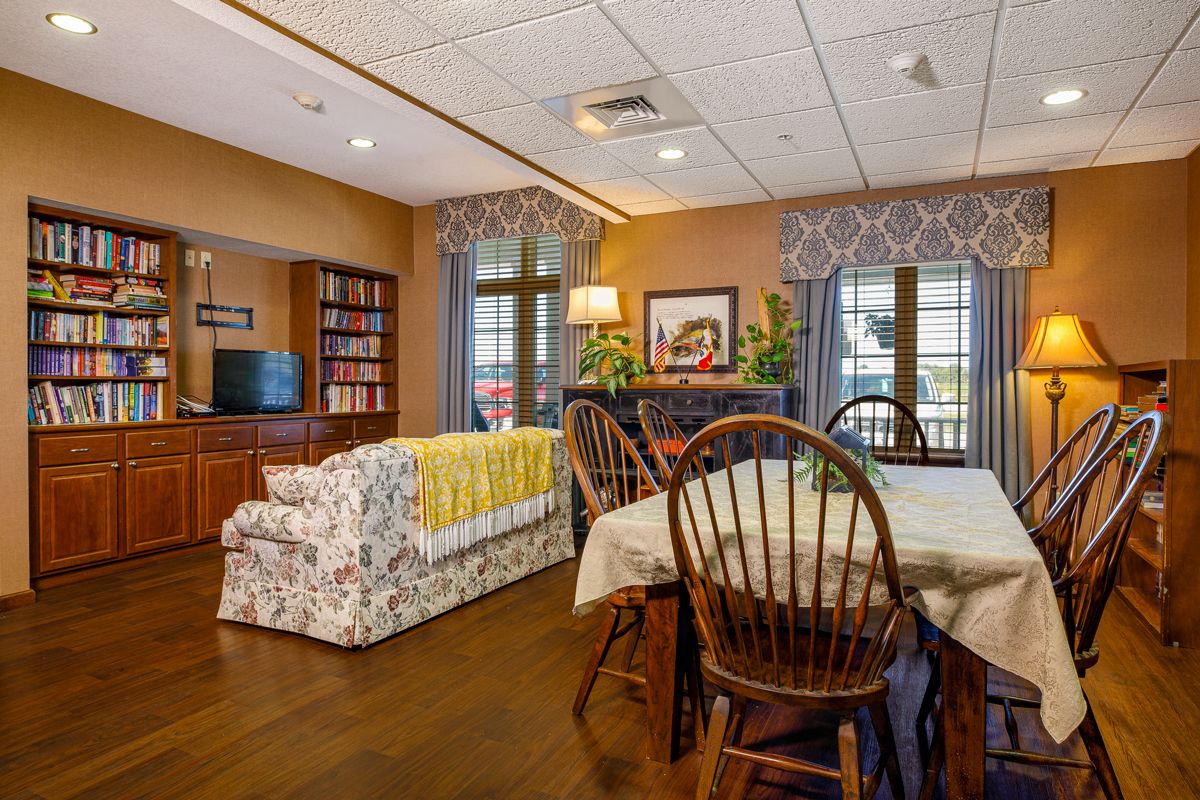 A cozy living room with a dining area, featuring a floral sofa, wooden dining table and chairs, bookshelves, and soft lighting.
