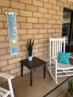 A cozy porch area with a welcome sign, rocking chairs, and a small table with a potted plant.