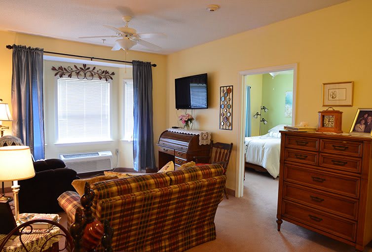 Cozy living room with a view into the bedroom, featuring a sofa, TV, and large windows with curtains.