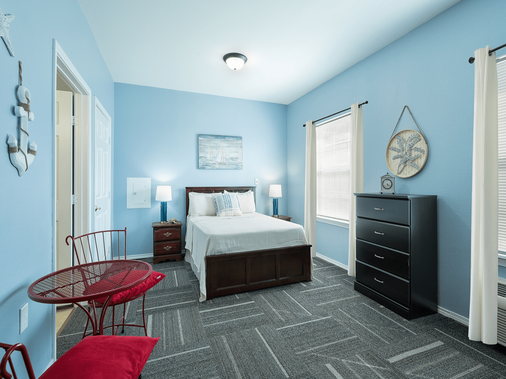 A modern bedroom with blue walls, featuring a bed, nightstands with lamps, a dark dresser, and a small table with red chairs.