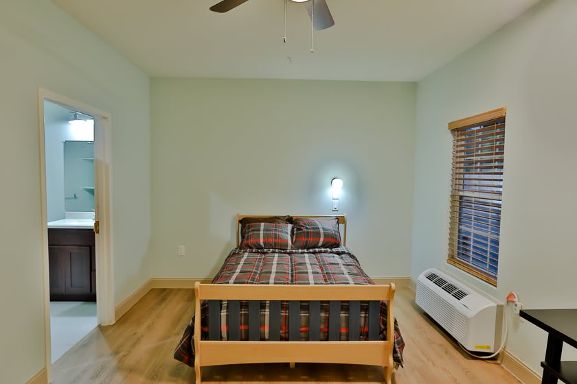 A minimalist bedroom featuring a single bed with plaid bedding, wooden floors, and a ceiling fan.