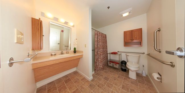 A spacious bathroom featuring a sink with a mirror, a toilet, and a shower area with a curtain. The room has tiled flooring and wall-mounted cabinets.