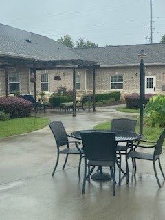 A patio area with tables and chairs, surrounded by a garden and buildings.